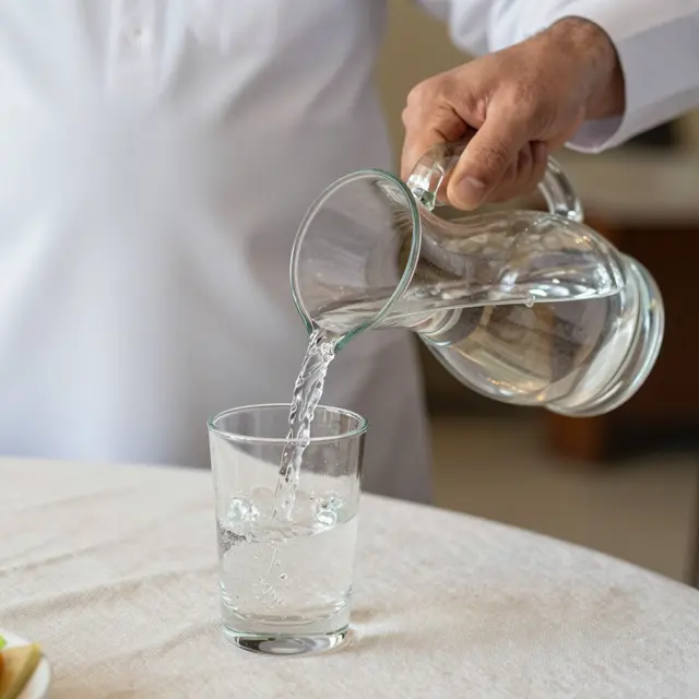A hand pours clear water from a glass carafe into a drinking glass on a linen tablecloth