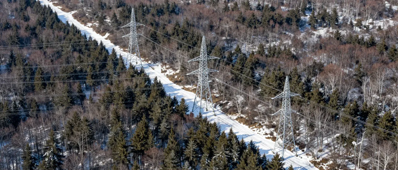 Aerial view of high-voltage power pylons along a snow-covered clearing through a winter forest