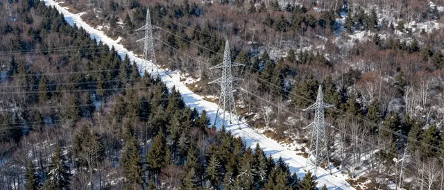 Aerial view of high-voltage power pylons along a snow-covered clearing through a winter forest