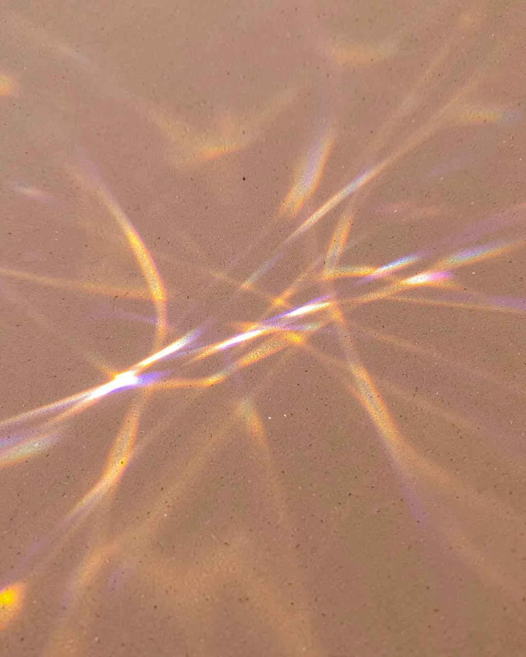 Iridescent gold and violet prismatic light streaks spread across a smooth sand-toned matte surface