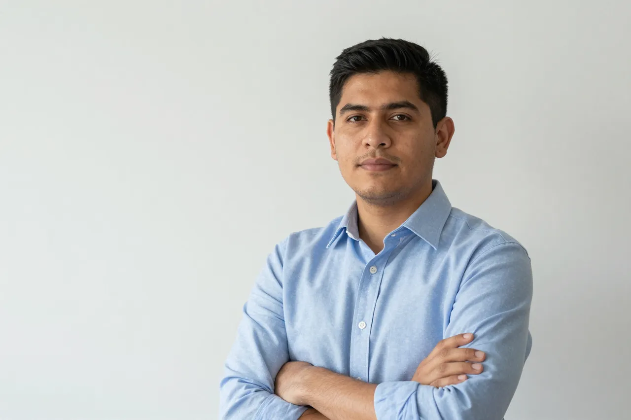 A Latino man in his thirties in a light blue shirt stands with arms folded against a plain white backdrop.