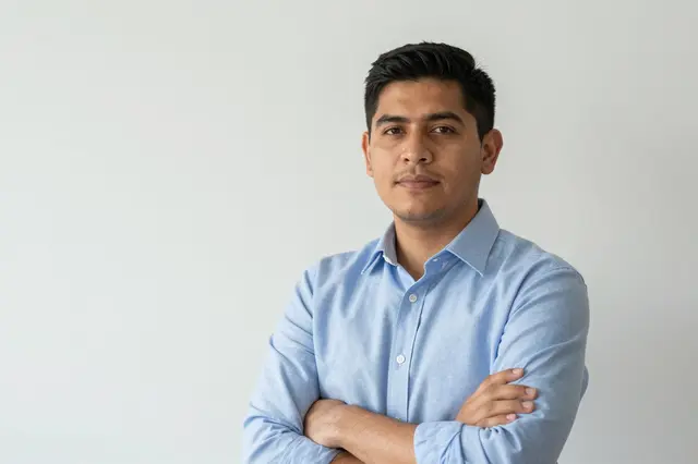 A Latino man in his thirties in a light blue shirt stands with arms folded against a plain white backdrop.