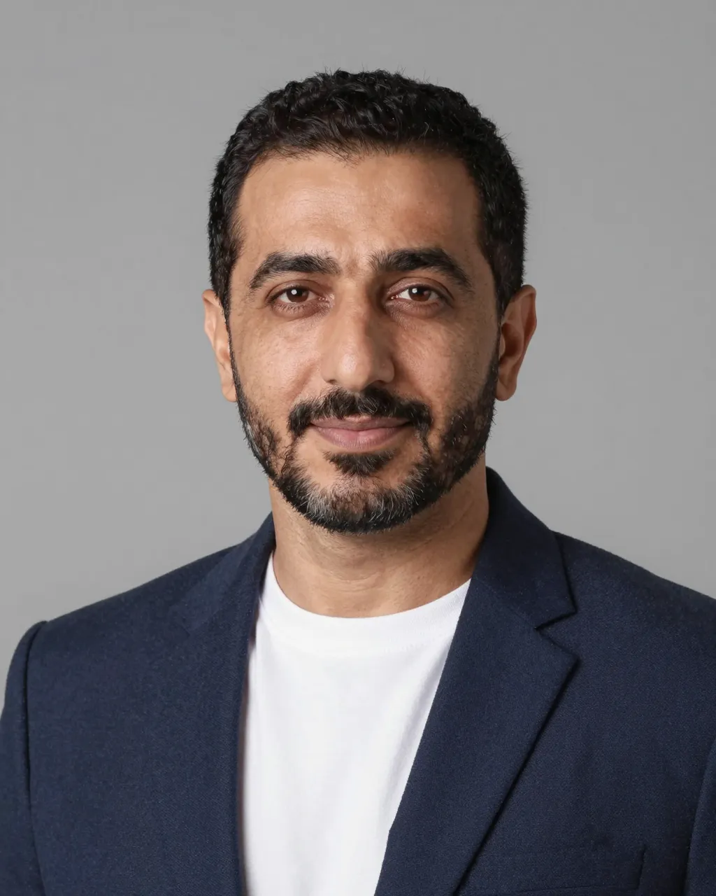 Studio headshot of a forties Middle Eastern man in a navy blazer over a white t-shirt against a gray backdrop