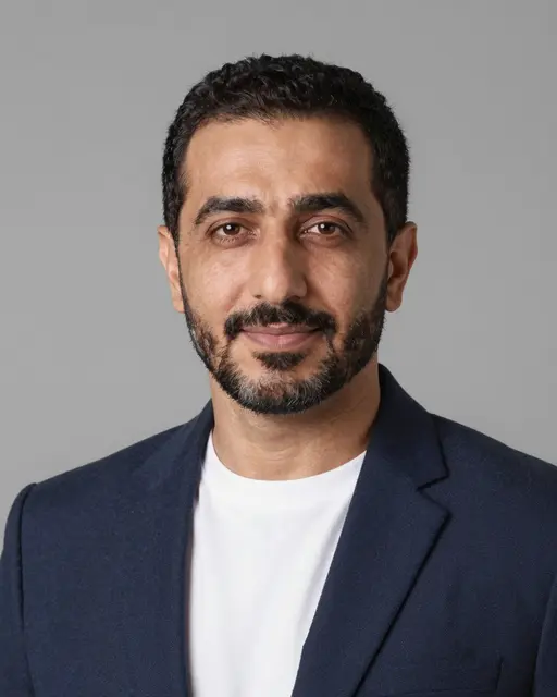 Studio headshot of a forties Middle Eastern man in a navy blazer over a white t-shirt against a gray backdrop