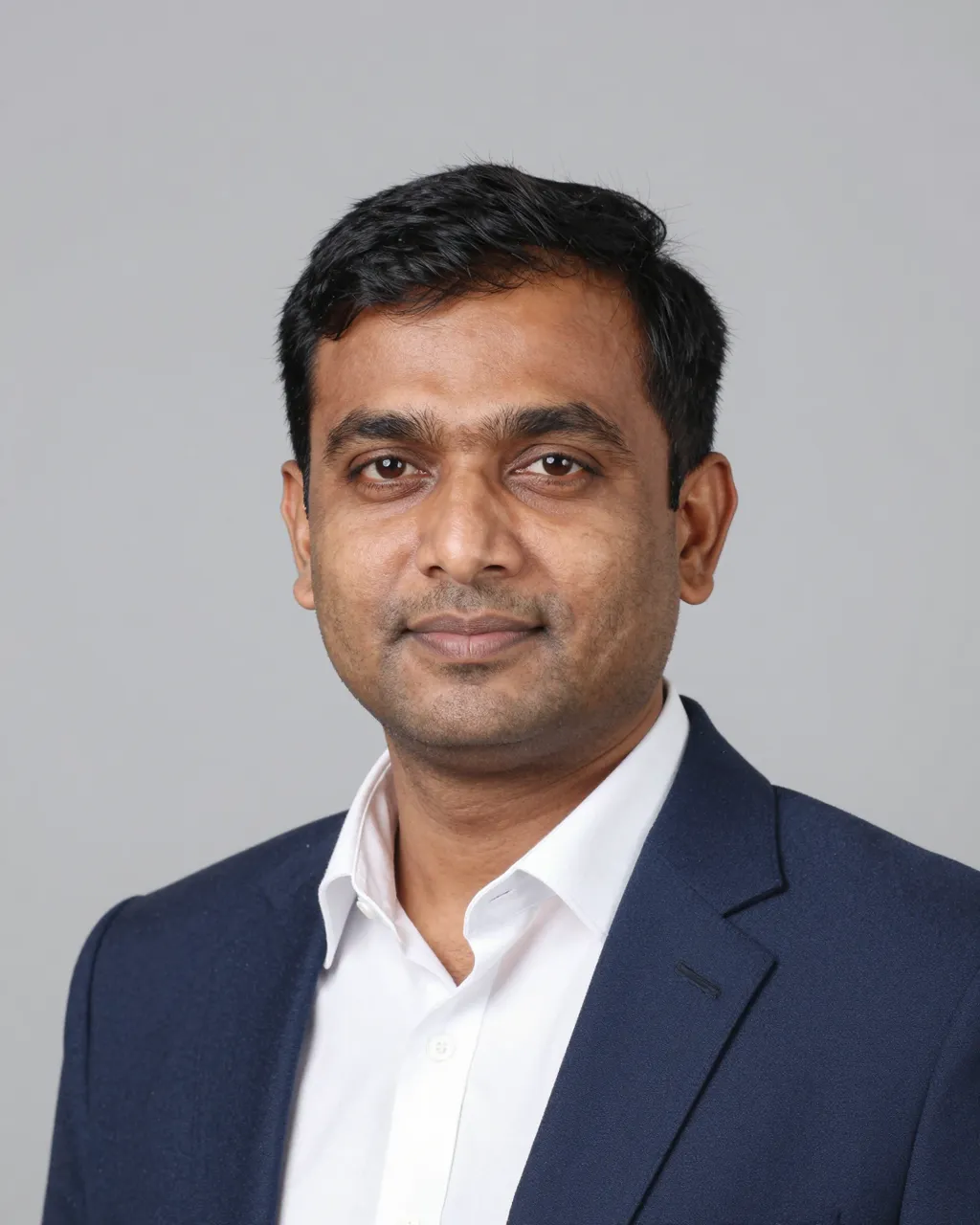 Head and shoulders portrait of a South Asian man in a navy blazer and white shirt against a plain gray backdrop