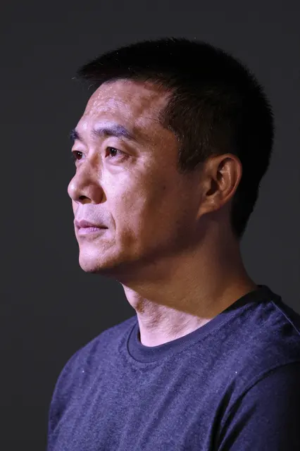 Profile view of a middle-aged East Asian man with faint violet lighting on his face against a dark background