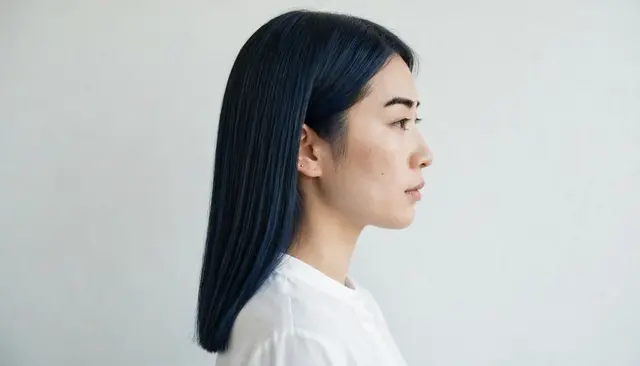 Side profile portrait of an East Asian woman with long blunt blue-black hair wearing a white blouse against a pale neutral