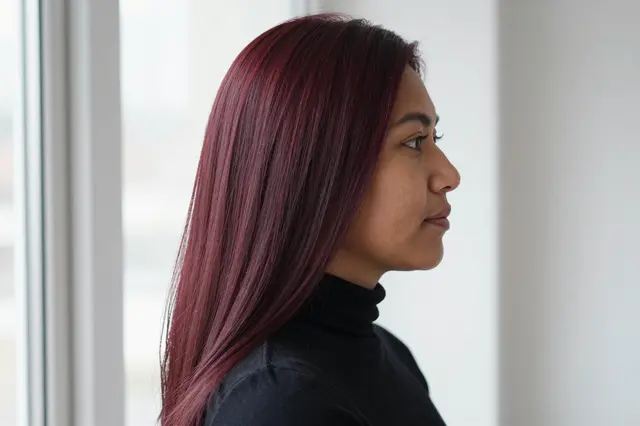 Side profile portrait of a woman with long straight burgundy hair wearing a black turtleneck near a bright window