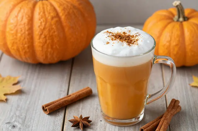 Cream-topped pumpkin spice latte sits with pumpkins, cinnamon sticks and fall leaves on a wooden table.