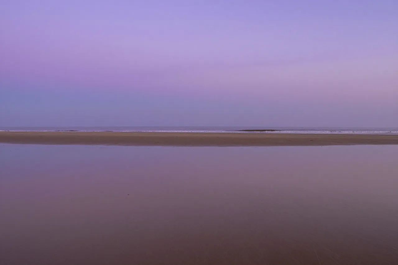 A calm flat beach at dusk, with soft purple sky mirrored in smooth wet sand