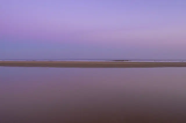 A calm flat beach at dusk, with soft purple sky mirrored in smooth wet sand