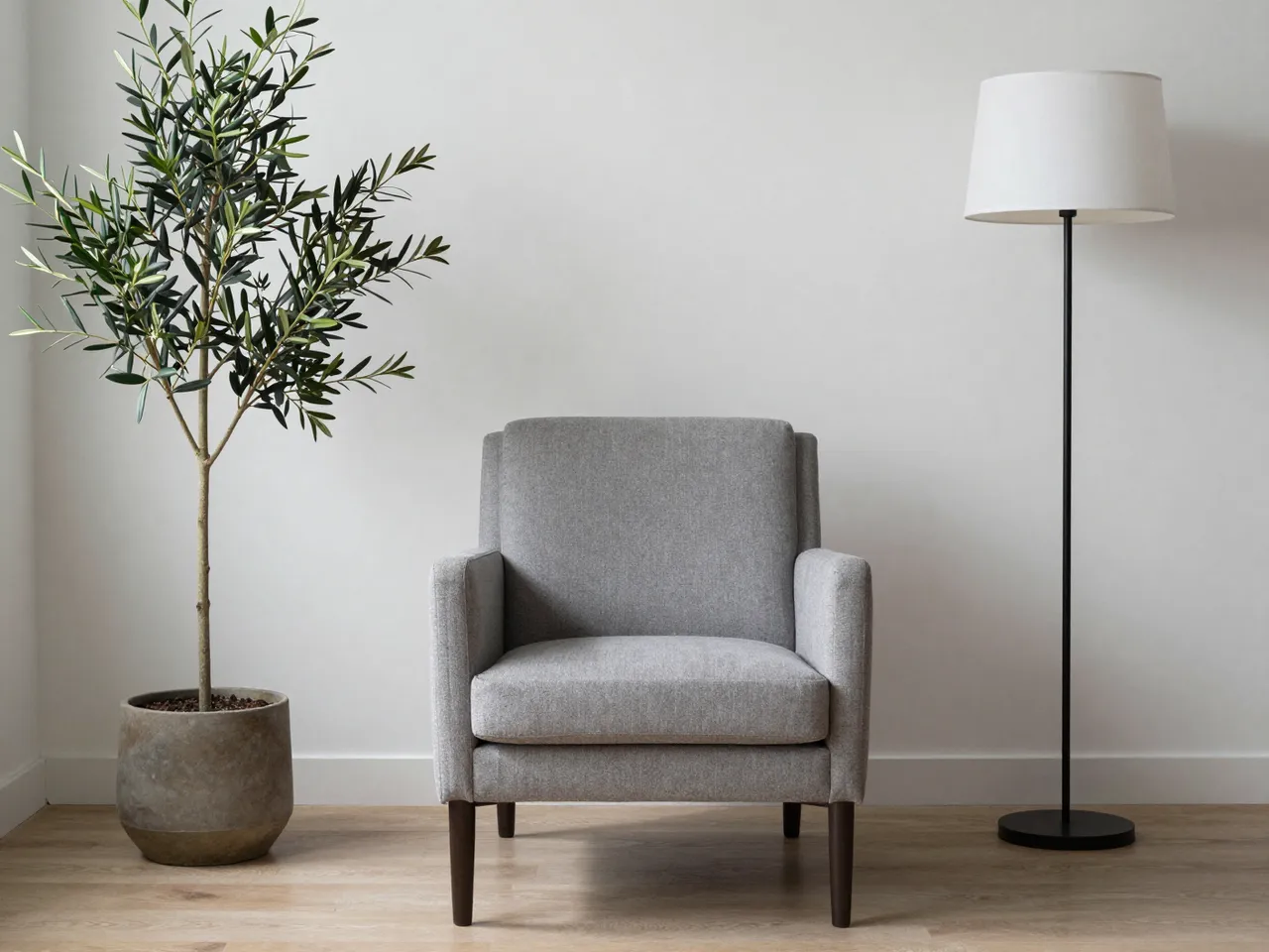 Gray upholstered armchair sits between a potted olive tree and a floor lamp against a plain light wall