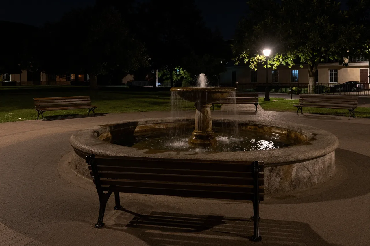 Illuminated stone fountain sits at the center of an empty paved town square lined with benches under a street lamp after dark