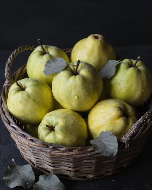 Fresh yellow-green quinces with gray leaves arranged in a woven wicker basket against a dark moody background