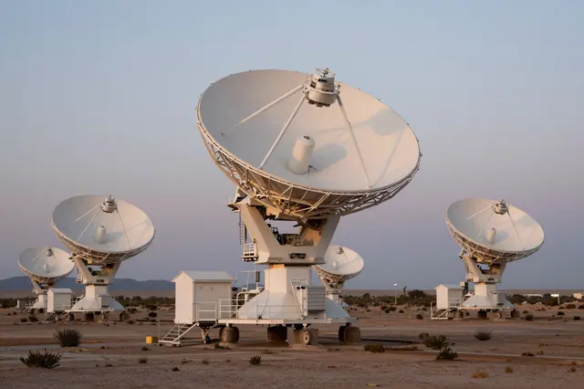 Array of white radio telescope dishes arranged across an arid desert plain at soft dusk