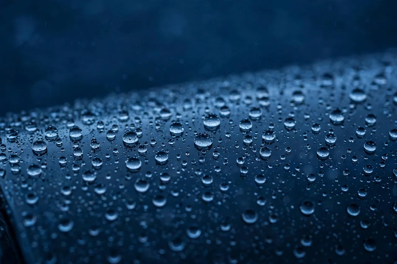 Sharp clear rain droplets beading on a dark blue glass surface against a soft blurred cool background