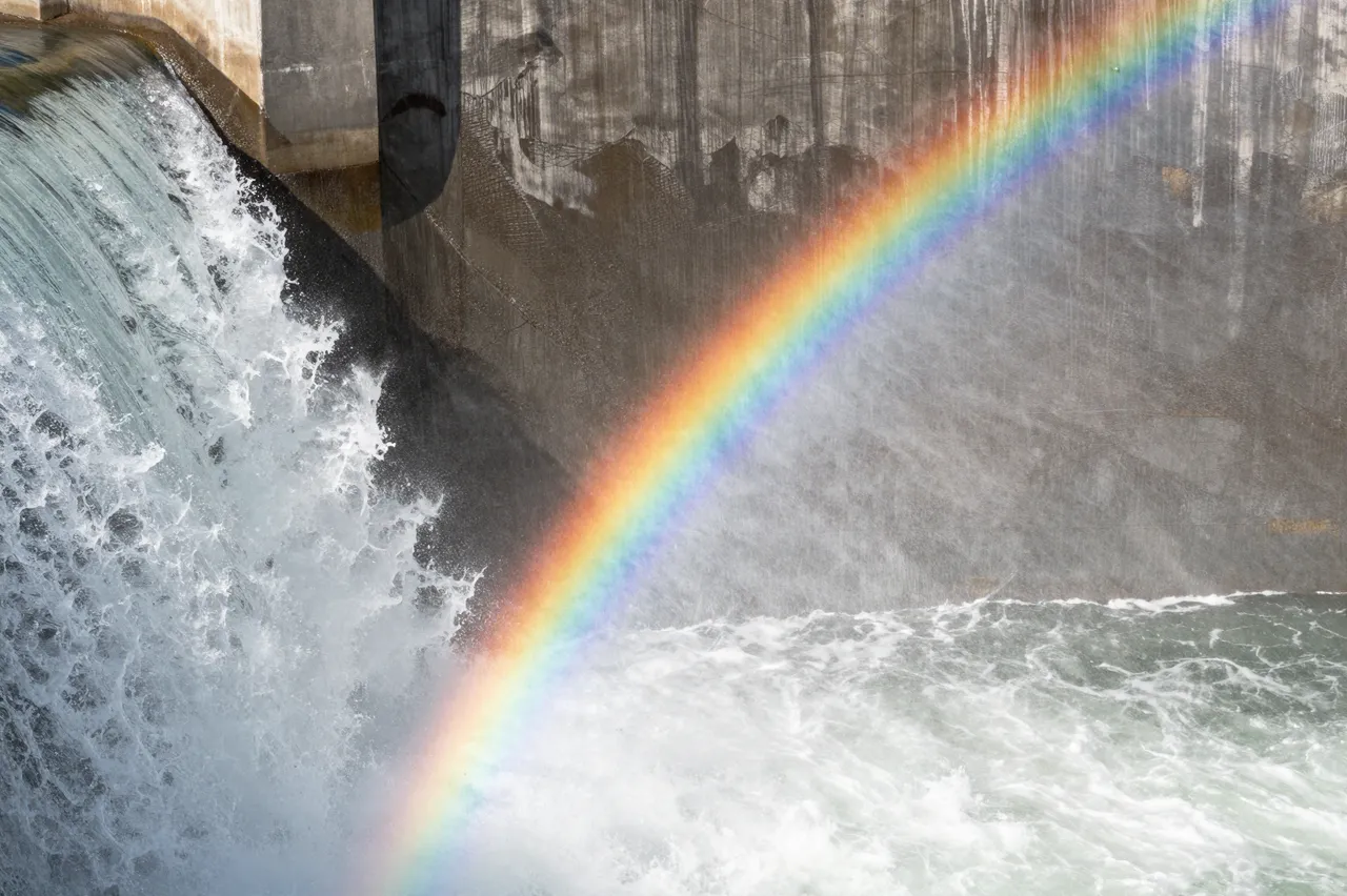 A diagonal rainbow stretches across churning white water flowing over a concrete dam spillway