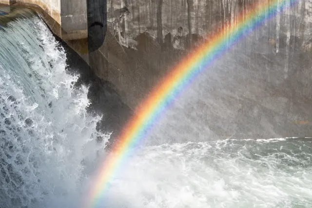 A diagonal rainbow stretches across churning white water flowing over a concrete dam spillway