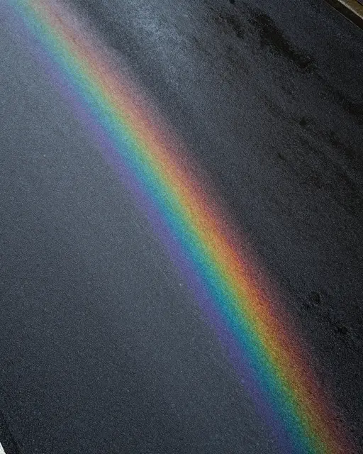 Top down view of wet dark asphalt with a diagonal iridescent rainbow oil sheen under overcast light