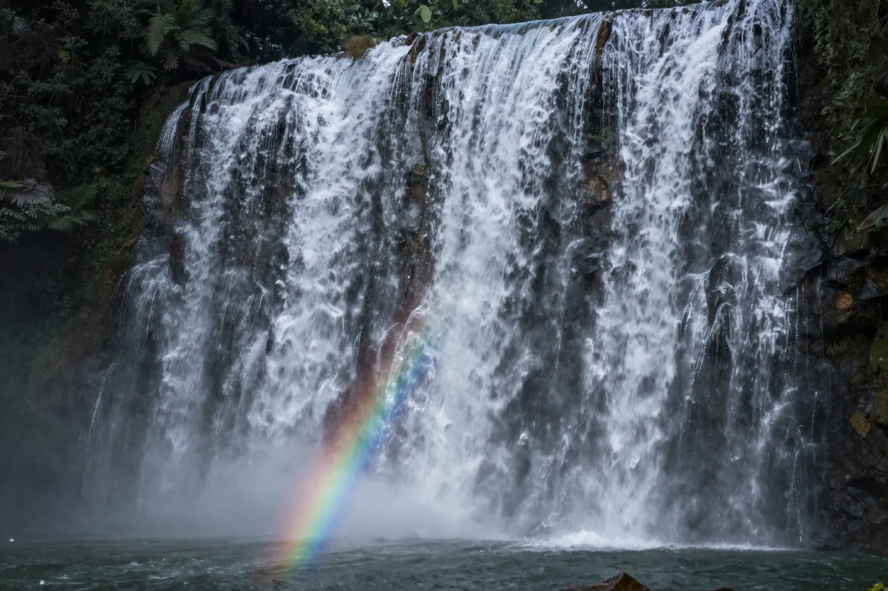 A broad cascading waterfall surrounded by dense jungle foliage has a bright rainbow arcing through its base mist.