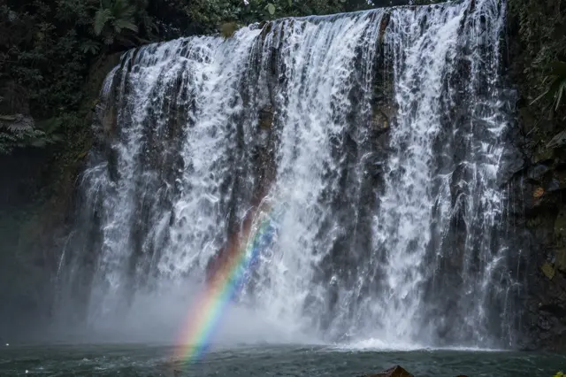 A broad cascading waterfall surrounded by dense jungle foliage has a bright rainbow arcing through its base mist.