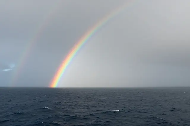 A bright primary rainbow and faint secondary rainbow arc down to the horizon over choppy dark ocean under a misty overcast