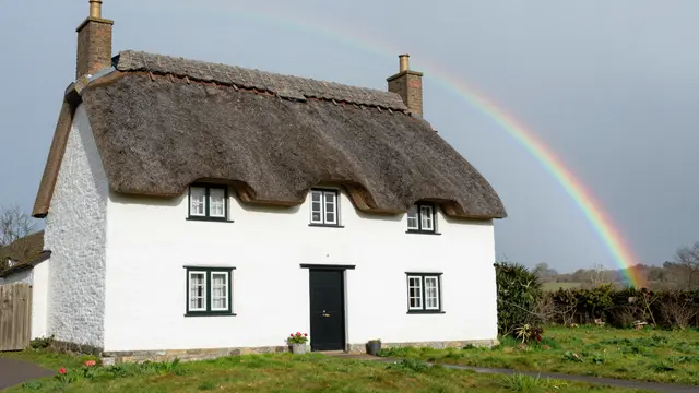 Whitewashed thatched stone cottage stands under a curved rainbow in green rural fields
