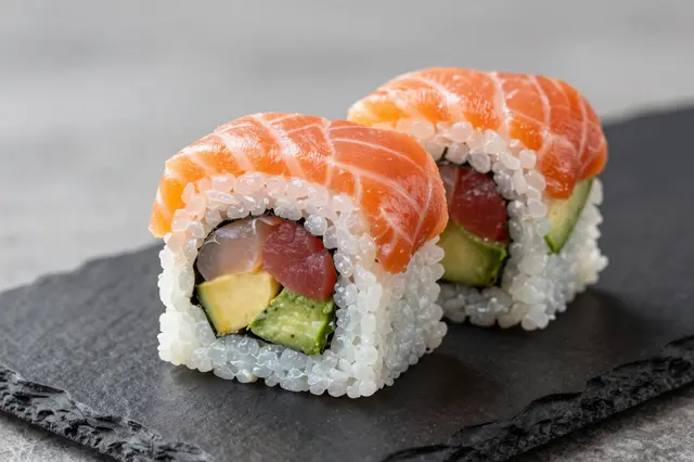 Two salmon-topped sushi roll pieces with tuna and avocado filling rest on a dark slate plate