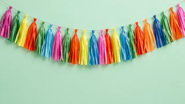 Swagged rainbow paper tassel garland hangs across a solid mint green wall with empty copy space below