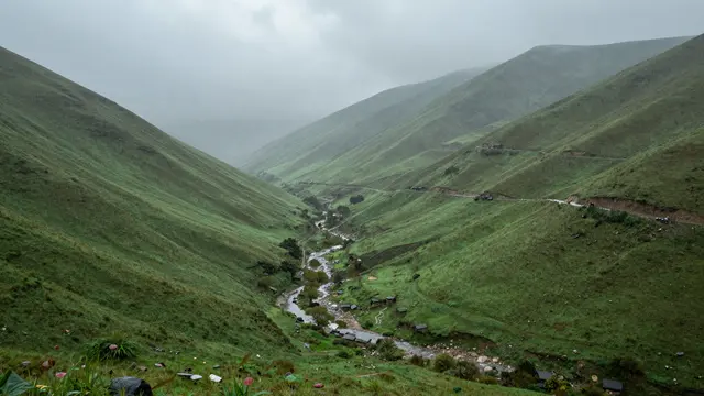 Overcast gray rain falls across a lush green mountain valley with a winding river and scattered small settlements.