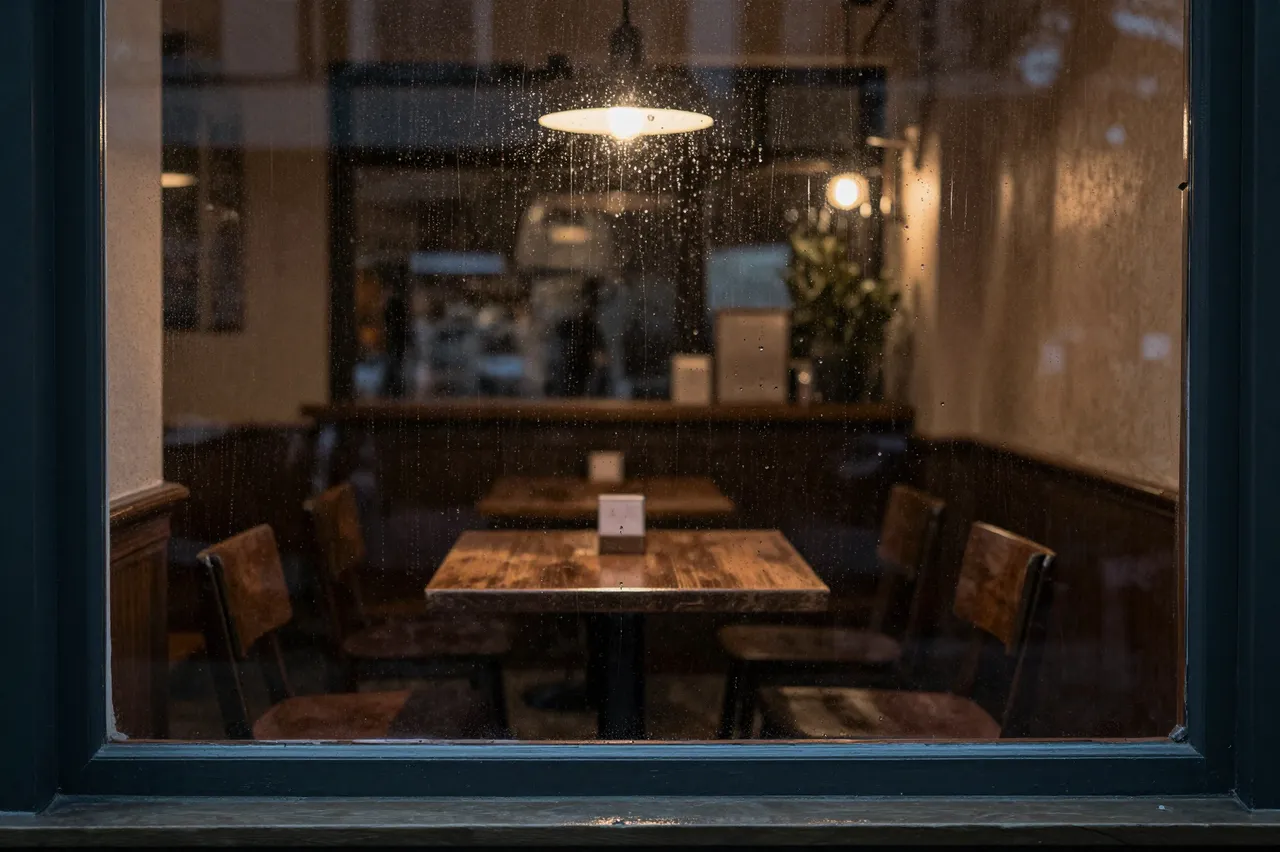 View through a rain-streaked cafe window showing empty wooden tables under warm pendant lights at night