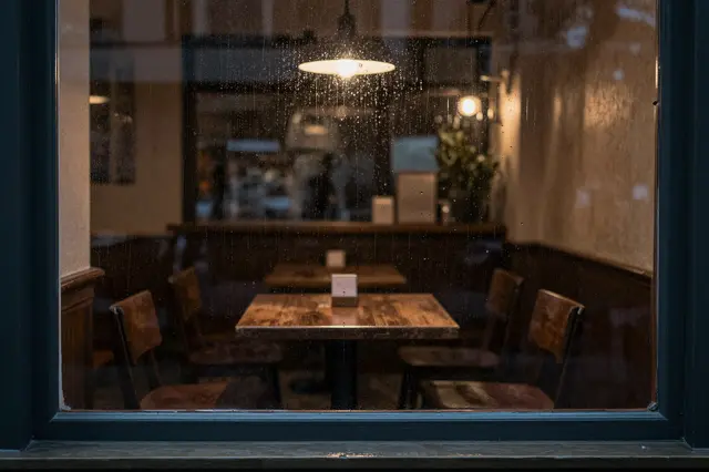 View through a rain-streaked cafe window showing empty wooden tables under warm pendant lights at night