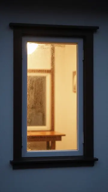 Warm golden interior light glows through a rain-streaked house window at night against a dark exterior wall
