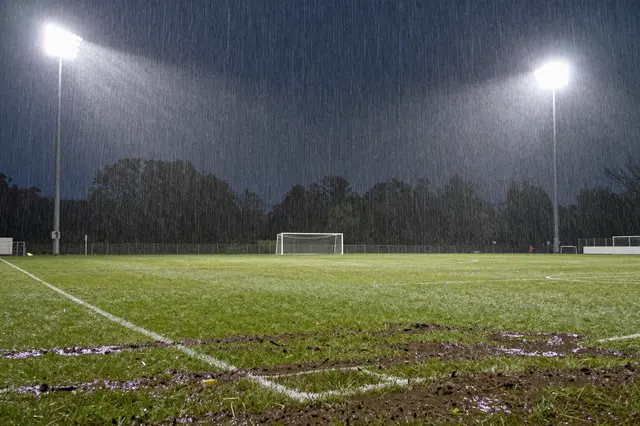 Floodlights illuminate falling rain over a muddy, wet soccer pitch at night