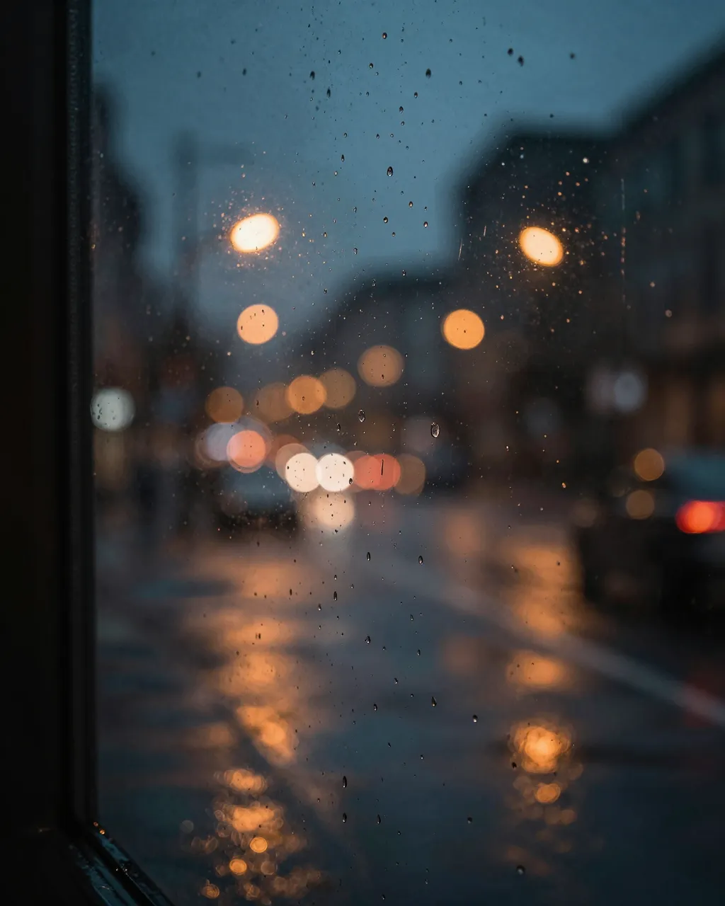 Rain droplets cling to a window overlooking a wet city street with warm blurred street light bokeh at dusk