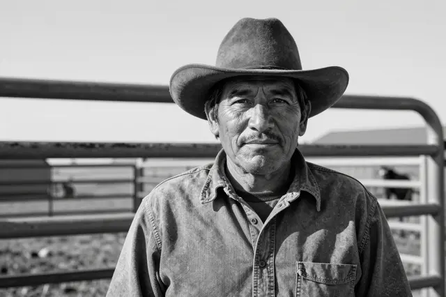Black and white close-up portrait of an older Indigenous rancher in a felt cowboy hat standing by a weathered metal corral