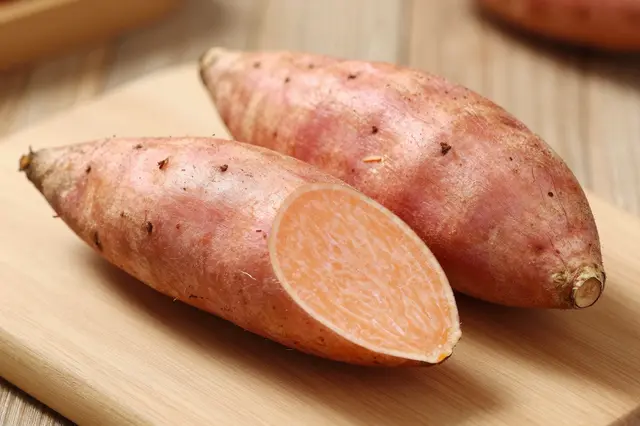 Whole and sliced raw sweet potatoes rest on a light wooden cutting board.