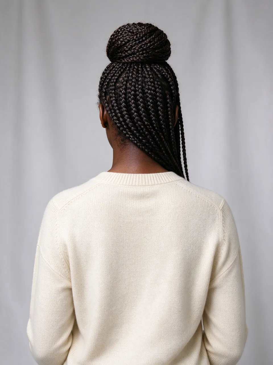 Back view of a Black woman with box braids in a high bun wearing a cream sweater in soft studio lighting