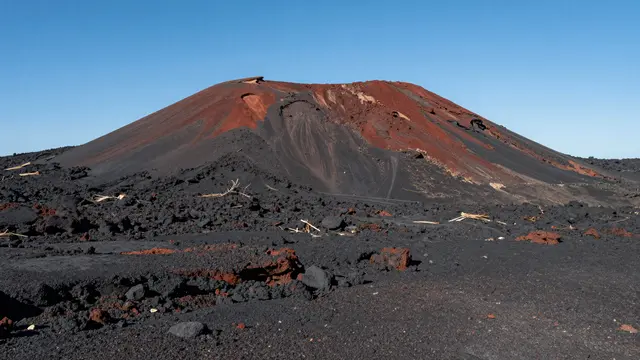A young red and black cinder cone volcano rises from a barren dark lava plain under a clear blue sky
