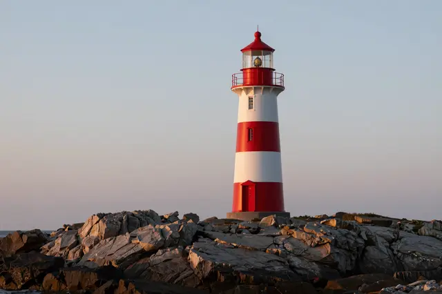A red and white striped lighthouse stands atop a rocky coastal point under a pale dusk sky