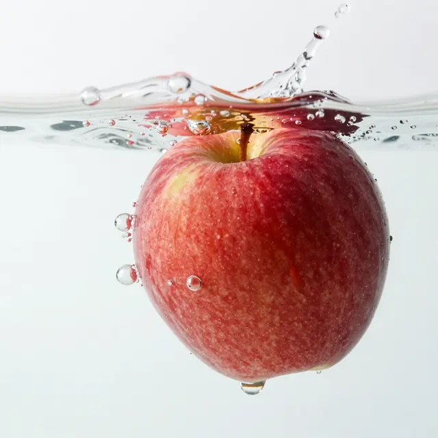 A red apple partially submerged in clear water with splashing droplets and rising bubbles against a bright white background