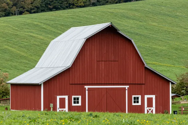 A classic red wooden barn with white trim and metal roof stands in a lush green rolling pasture