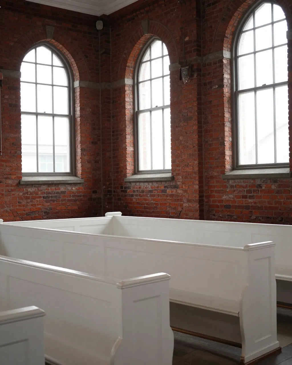 Empty church interior with red brick walls, arched windows and rows of white painted pews.