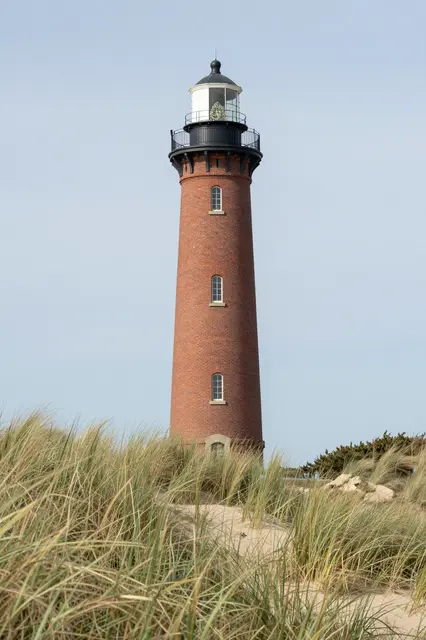 Tall red brick lighthouse stands among grassy coastal sand dunes under a pale clear sky