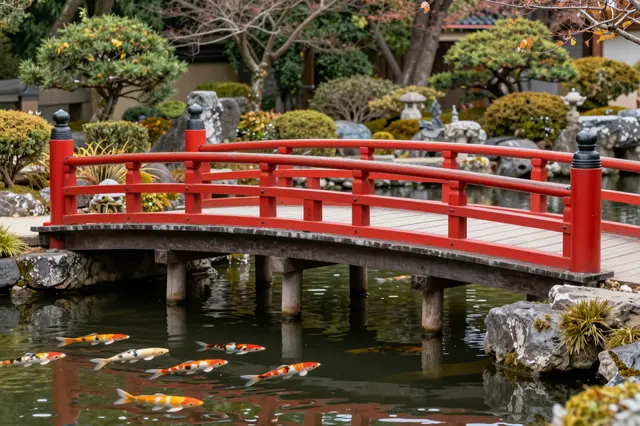A bright red wooden arched bridge spans a calm koi pond with swimming koi fish in a traditional Japanese garden.