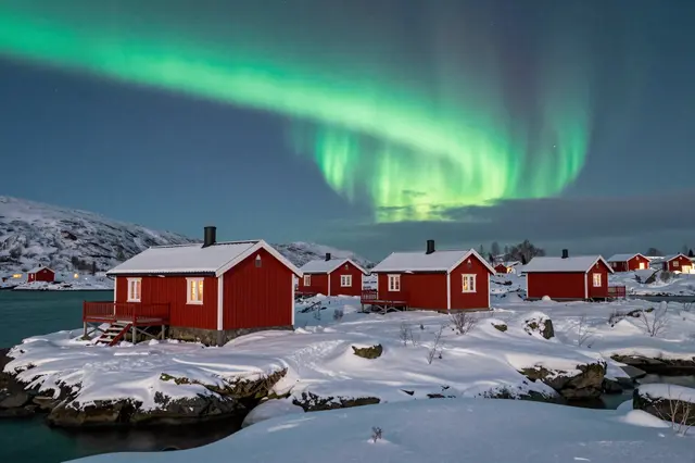 Glowing green northern lights arc over red wooden cabins on snow-covered rocky islets in arctic Norway