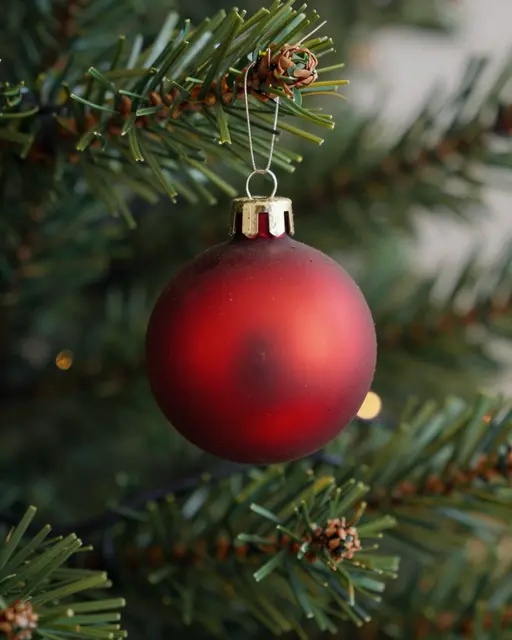 A matte red Christmas bauble hangs from a pine branch with soft golden bokeh lights in the background.