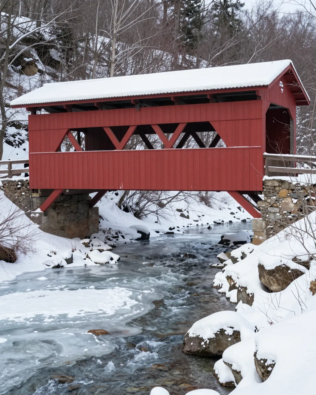 A red snow-covered wooden covered bridge spans a partially frozen winter creek surrounded by snow