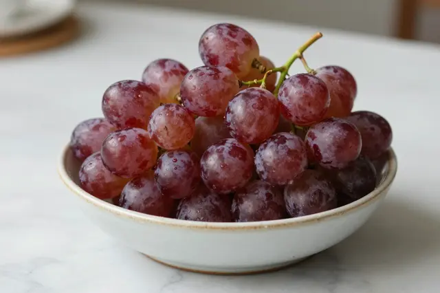 Bunch of fresh red grapes resting in a white ceramic bowl on a light marble countertop