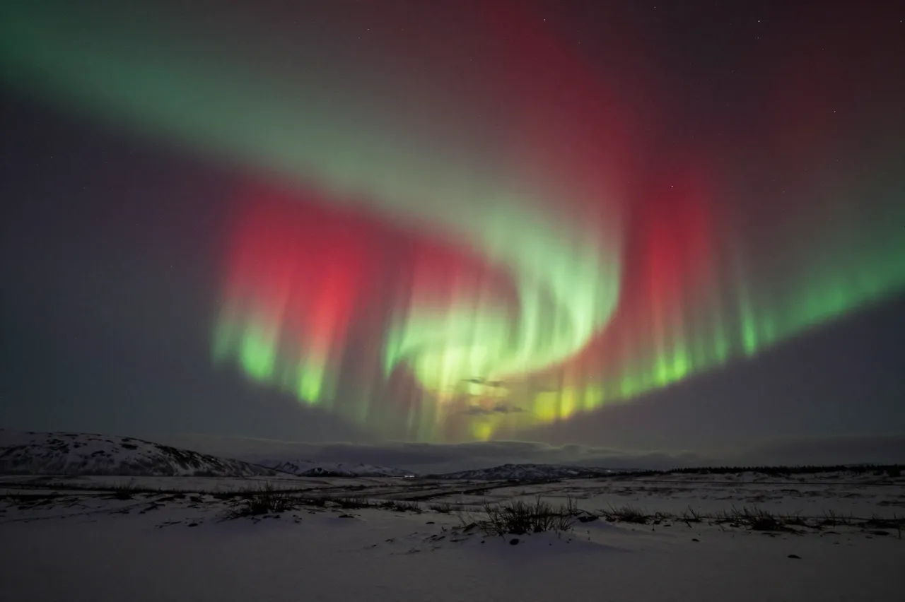 Vibrant red and green aurora arcs across a dark starry sky above a snow-covered northern tundra landscape.